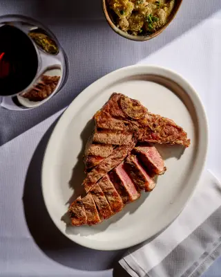 Carne grelhada fatiada em prato branco, vista de cima, com taça de vinho e acompanhamento